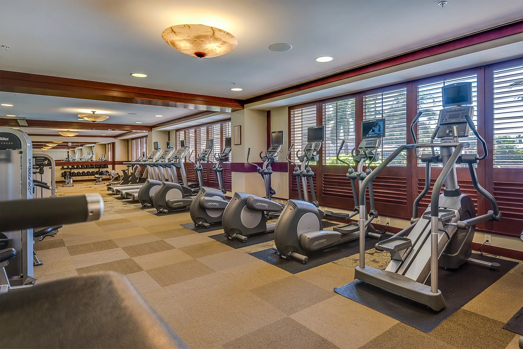 Un gimnasio con máquinas de cardio junto a grandes ventanas.