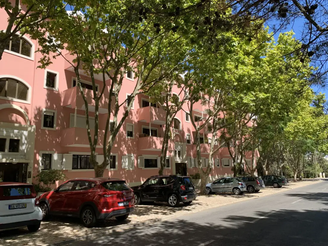 Un edificio de apartamentos rosa se encuentra junto a una calle arbolada con coches aparcados.