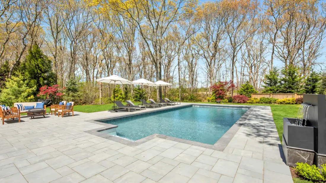 An outdoor swimming pool sits in a paved patio surrounded by trees.