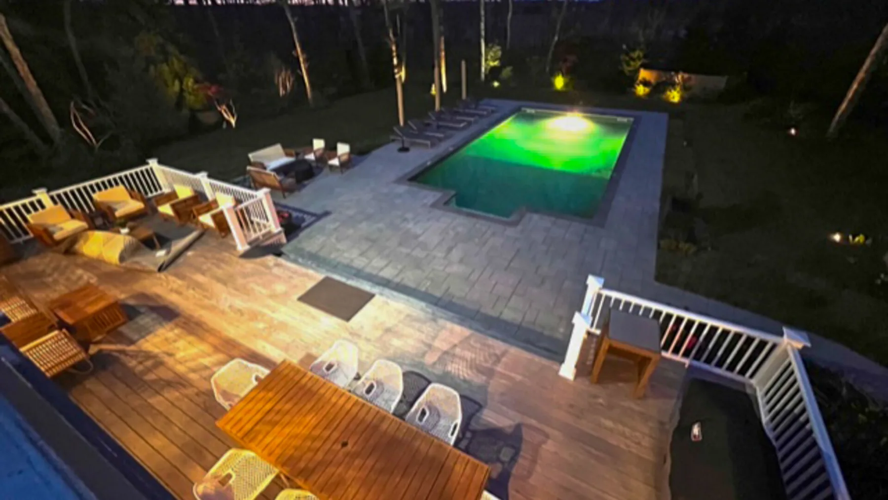An outdoor pool beside a wooden deck with seating, illuminated at night.