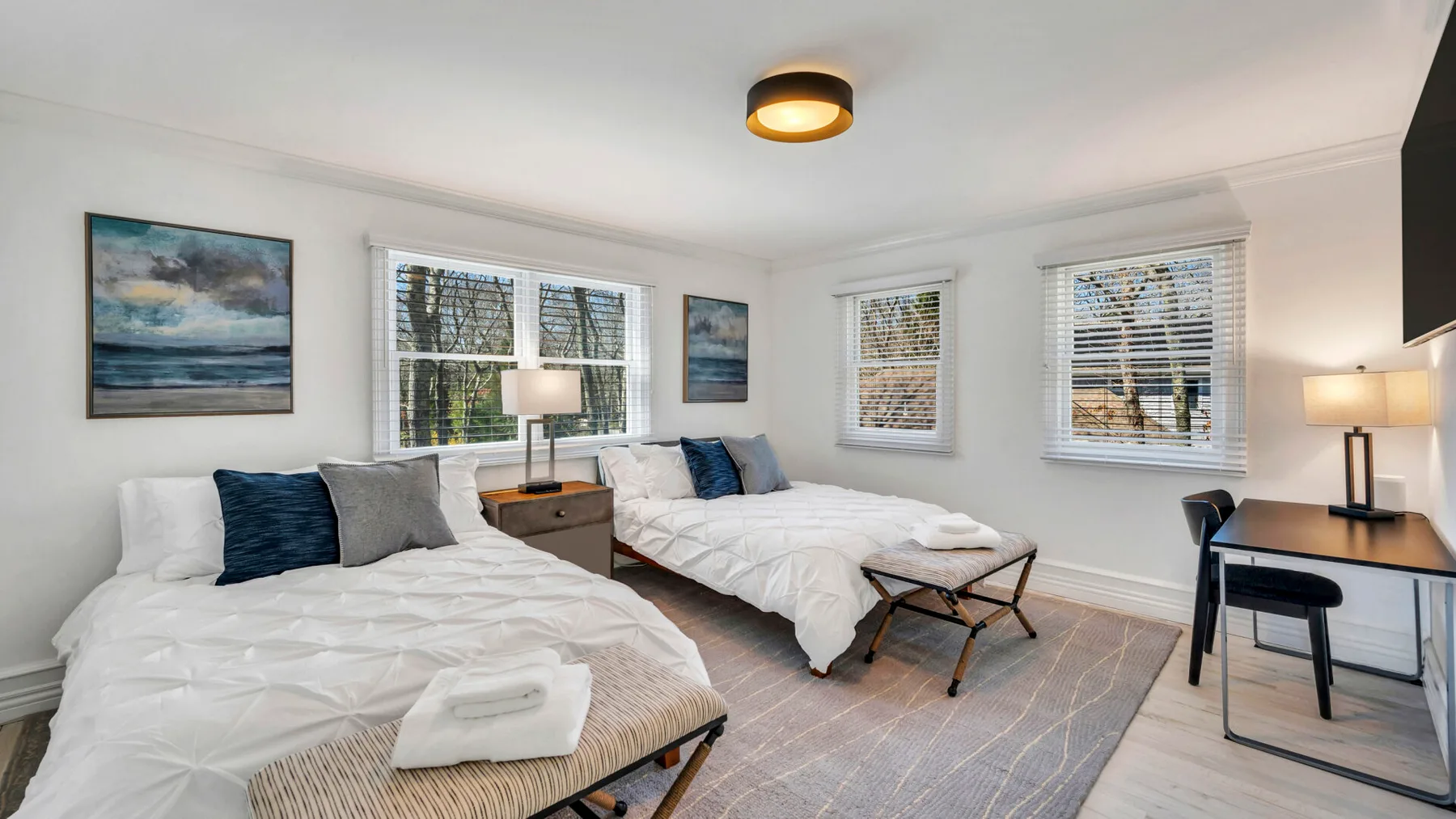 A bright bedroom with two beds, bedside tables, and large windows.