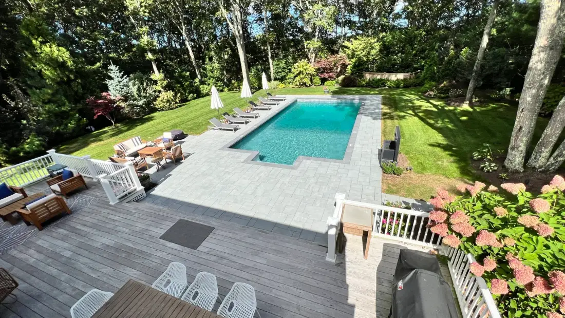 An outdoor pool and paved patio are surrounded by lawn and mature trees.
