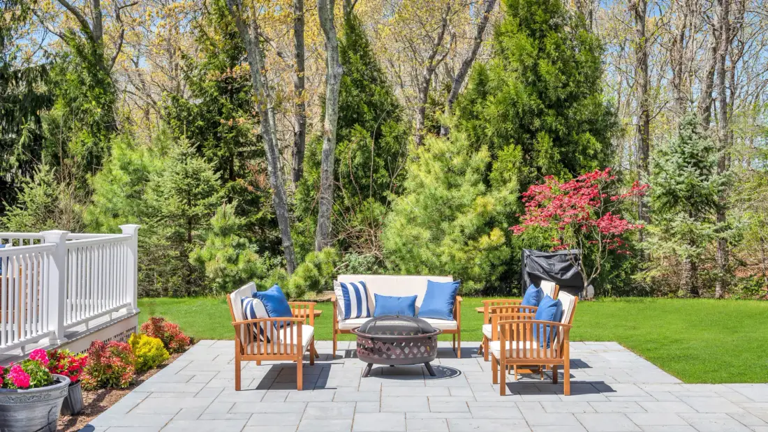 An outdoor patio has wooden seating around a fire pit, with trees and garden beds nearby.