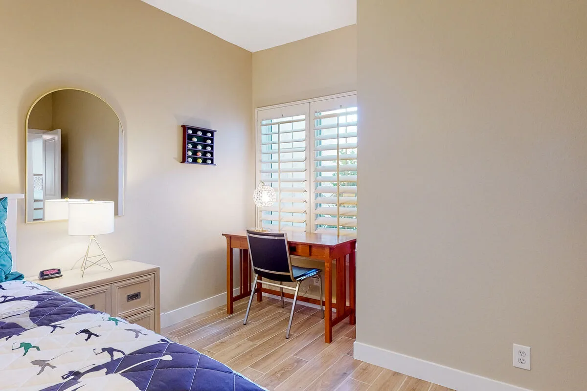 Un dormitorio con una cama junto a un escritorio y una silla cerca de ventanas con contraventanas.