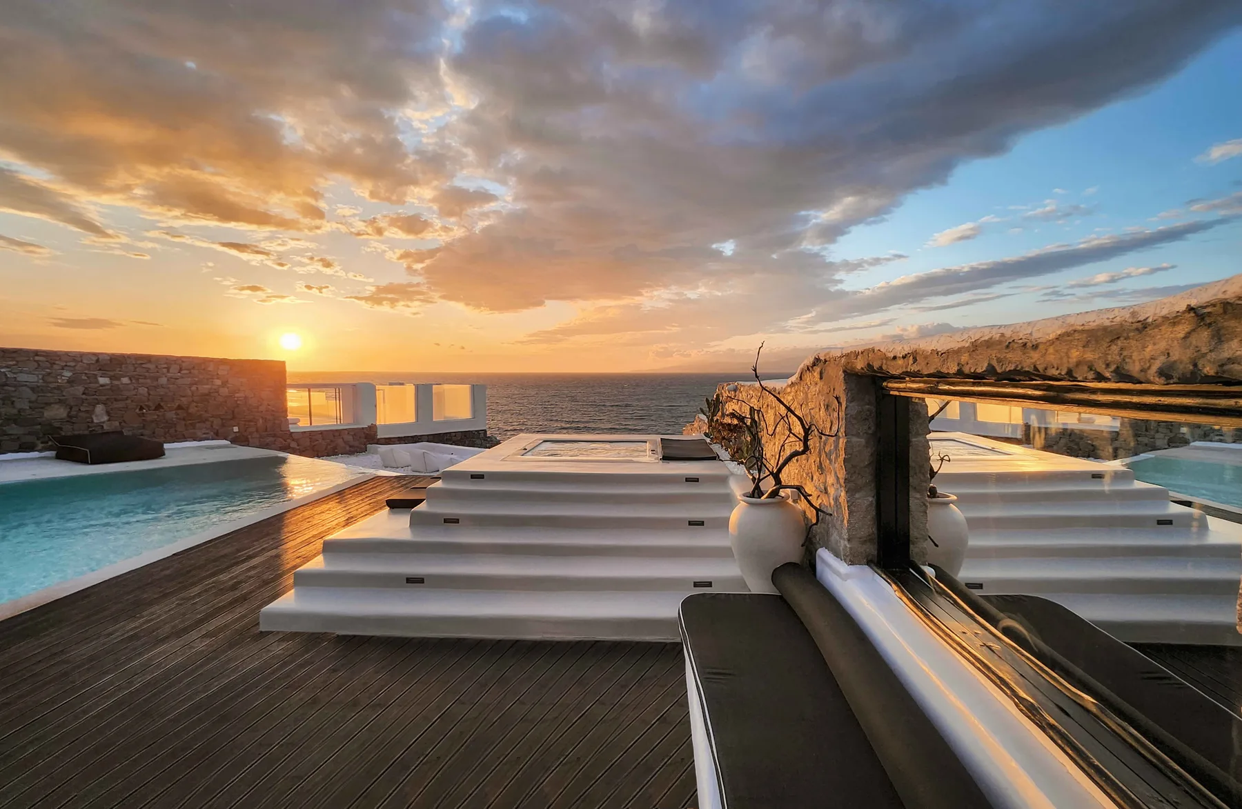 A rooftop pool terrace overlooks the sea at sunset, with white sun loungers.