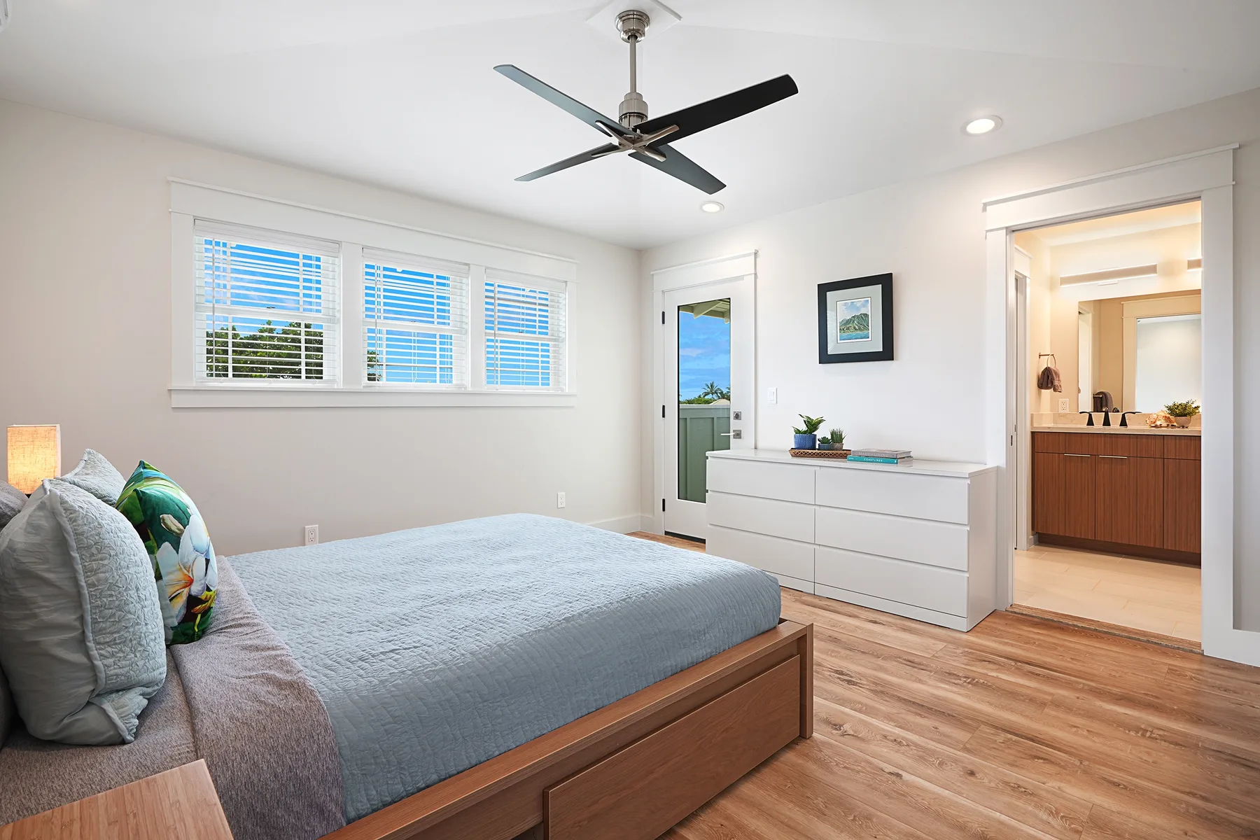 Un dormitorio luminoso con una cama, ventanas amplias y una cómoda blanca.
