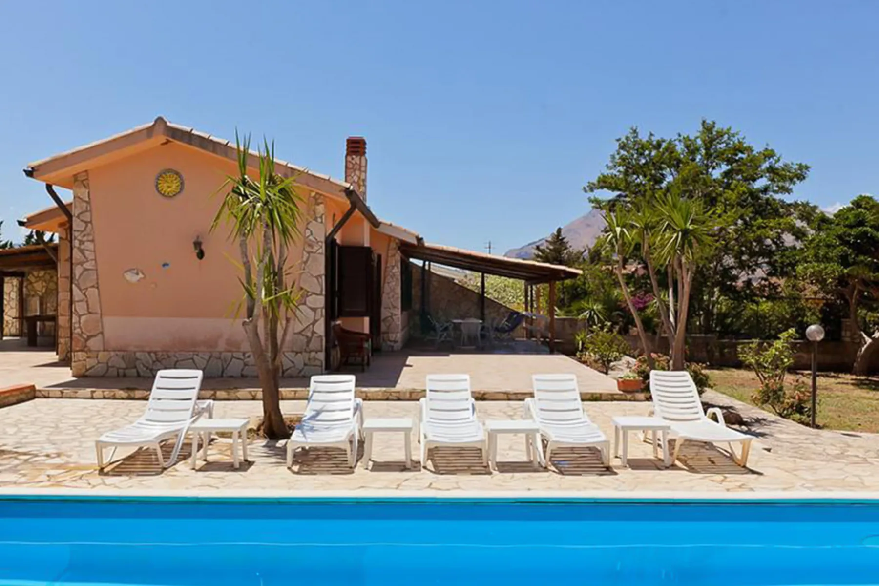 A poolside patio with lounge seating beside a stucco house and garden.