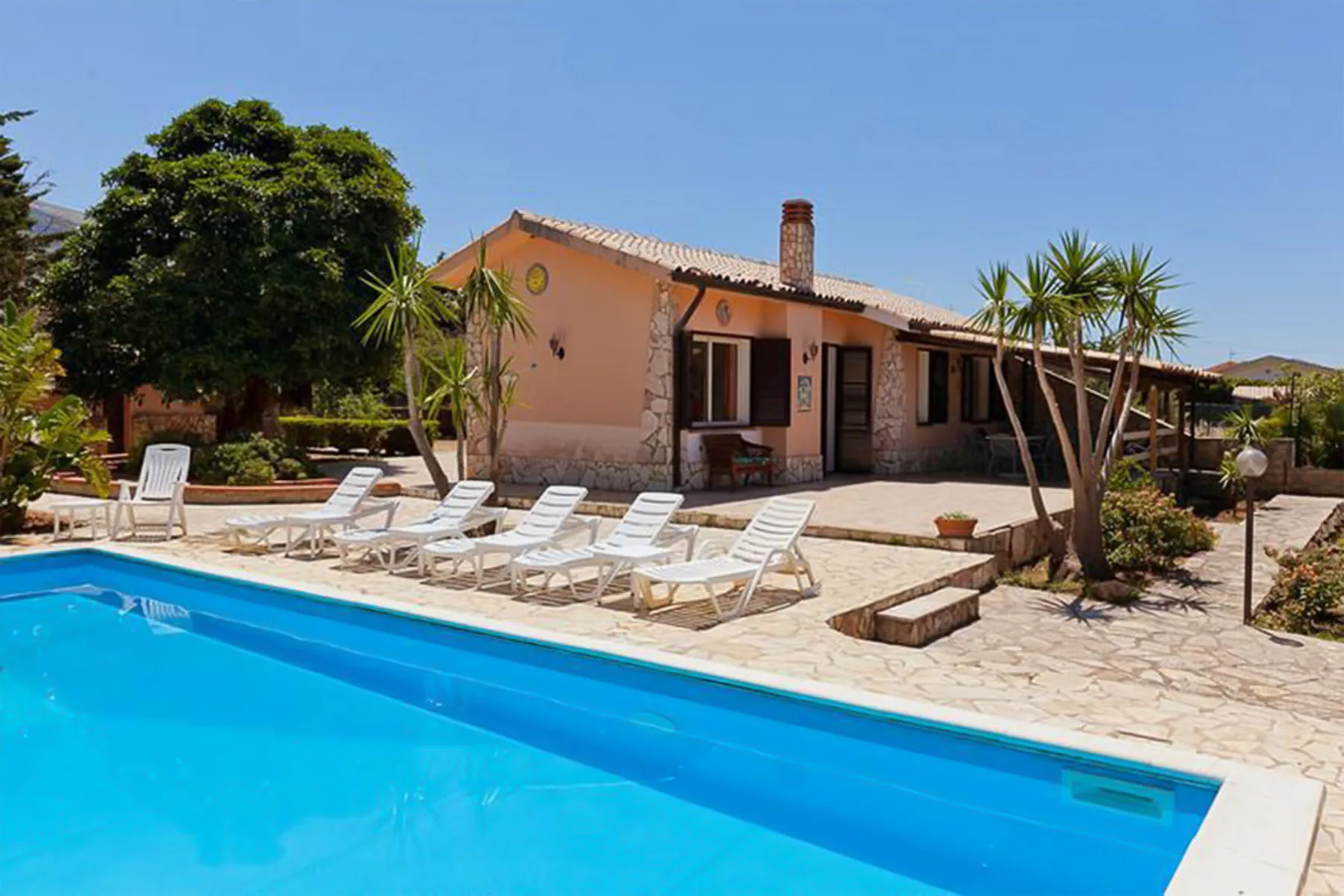 A swimming pool and lounge chairs sit in front of a single-story house.
