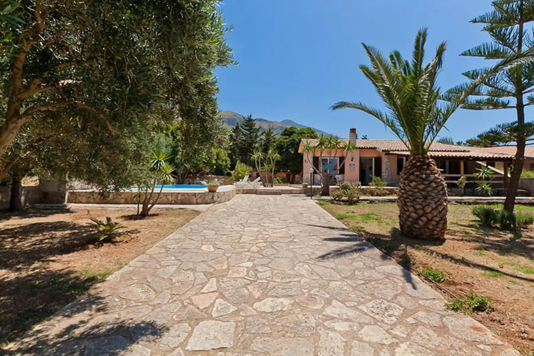A stone path leads through a landscaped yard toward a low house and pool.