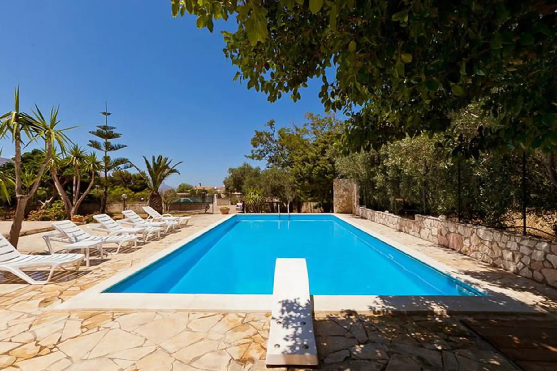An outdoor swimming pool is surrounded by stone paving, lounge seating, and trees.
