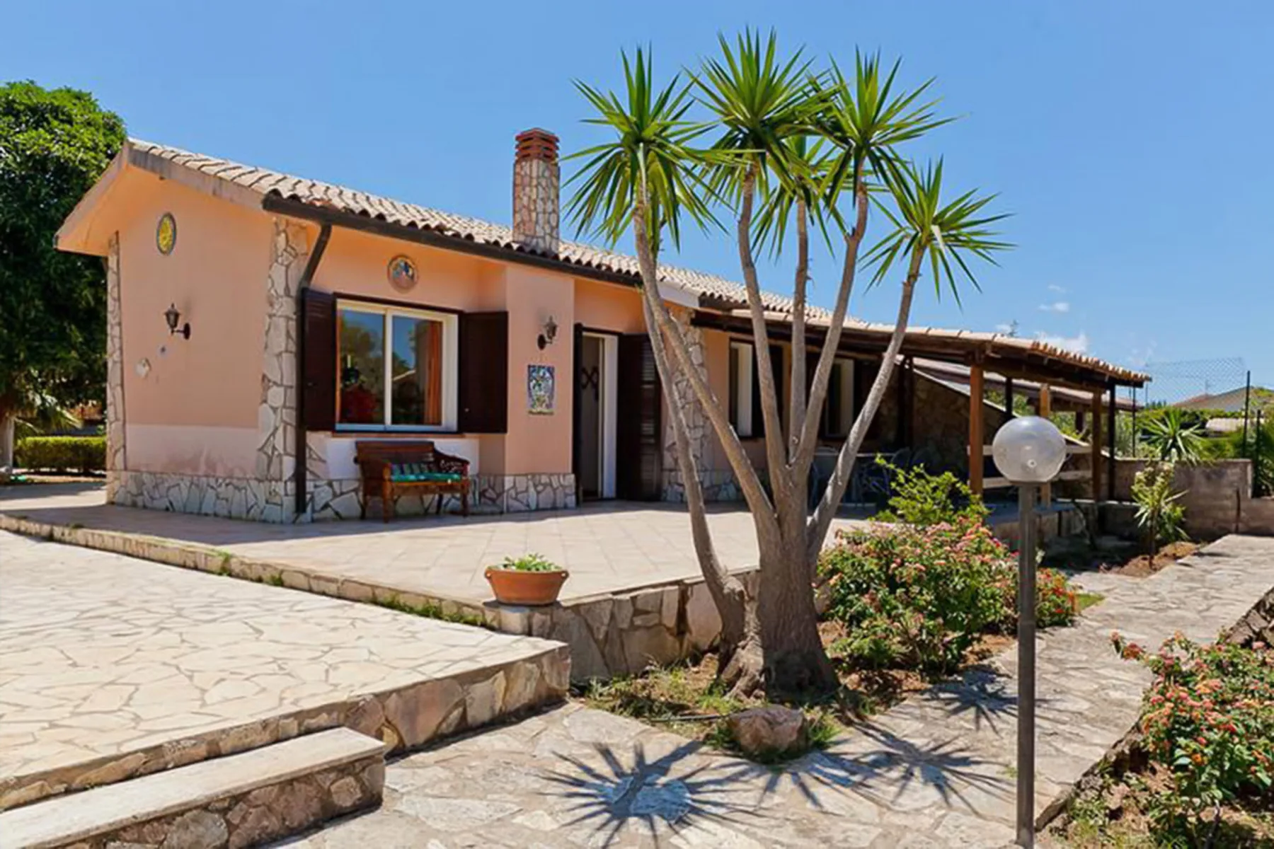 A single-story house with a paved patio, tall plants, and a covered outdoor area.