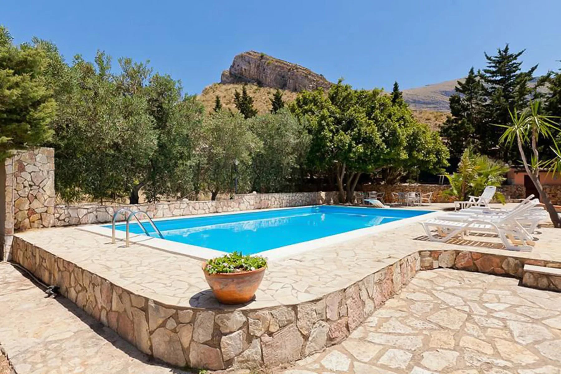 An outdoor swimming pool sits on a stone patio among trees and lounge chairs.