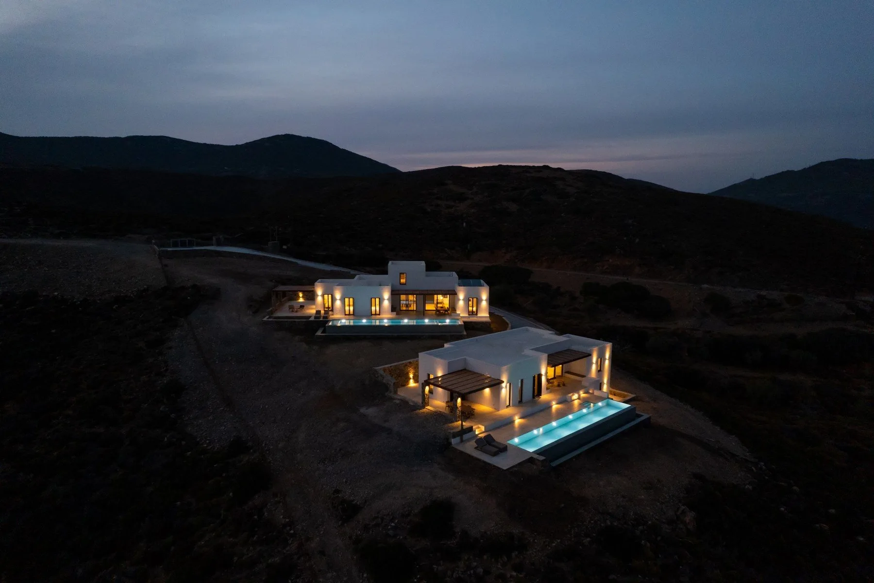 Aerial view of two illuminated villas beside swimming pools in a dark hillside landscape.