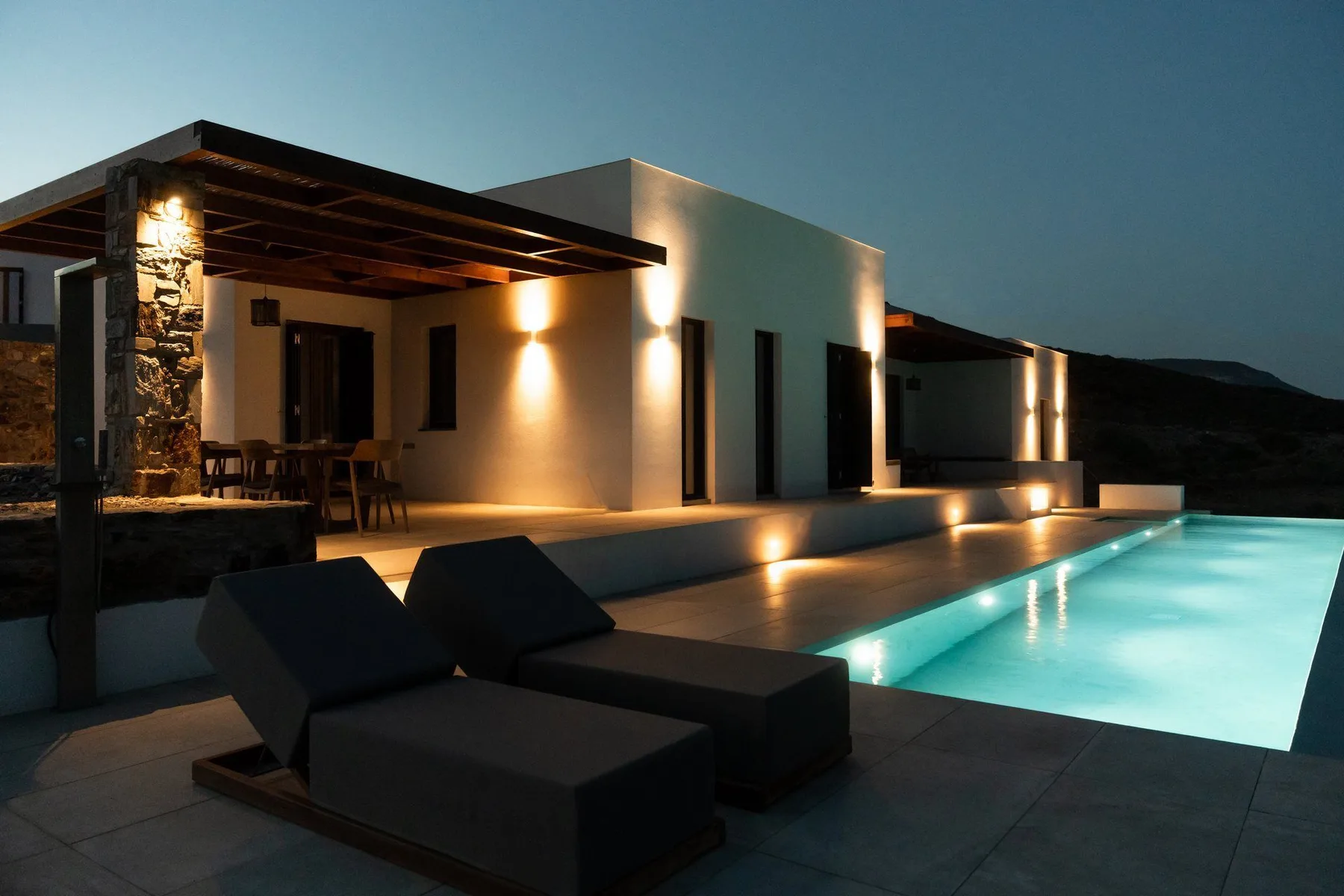 A modern house patio with lounge seating beside a lit swimming pool.