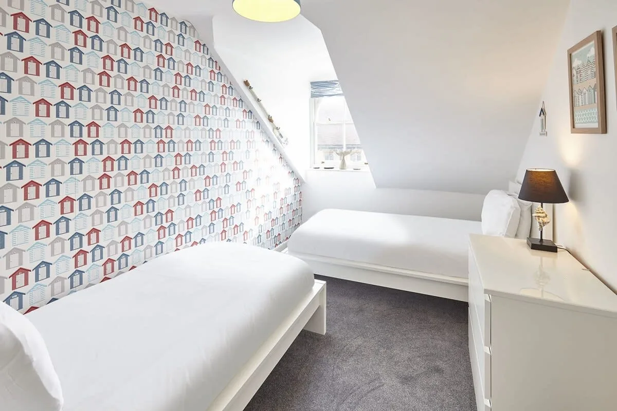 A bright bedroom with two beds and patterned wallpaper.