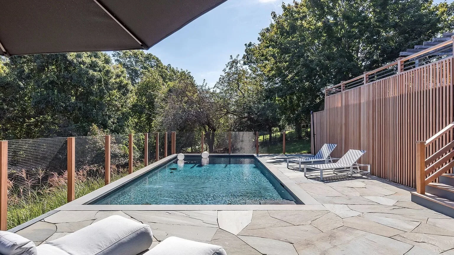 Una piscina al aire libre está junto a un patio pavimentado con tumbonas y árboles.