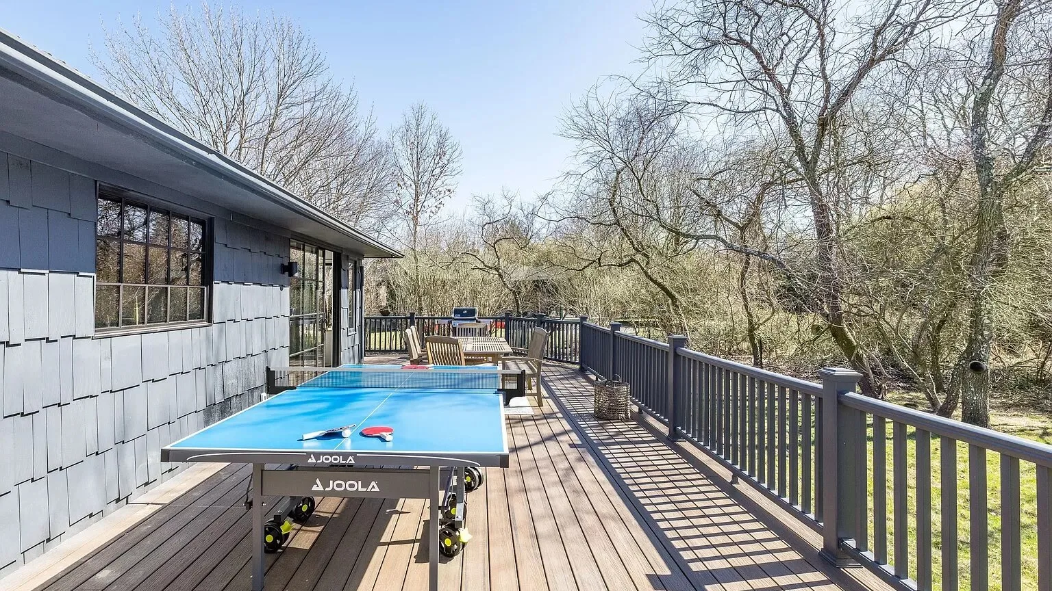 Una terraza junto a un edificio de revestimiento gris tiene una mesa de ping-pong azul y una baranda oscura.