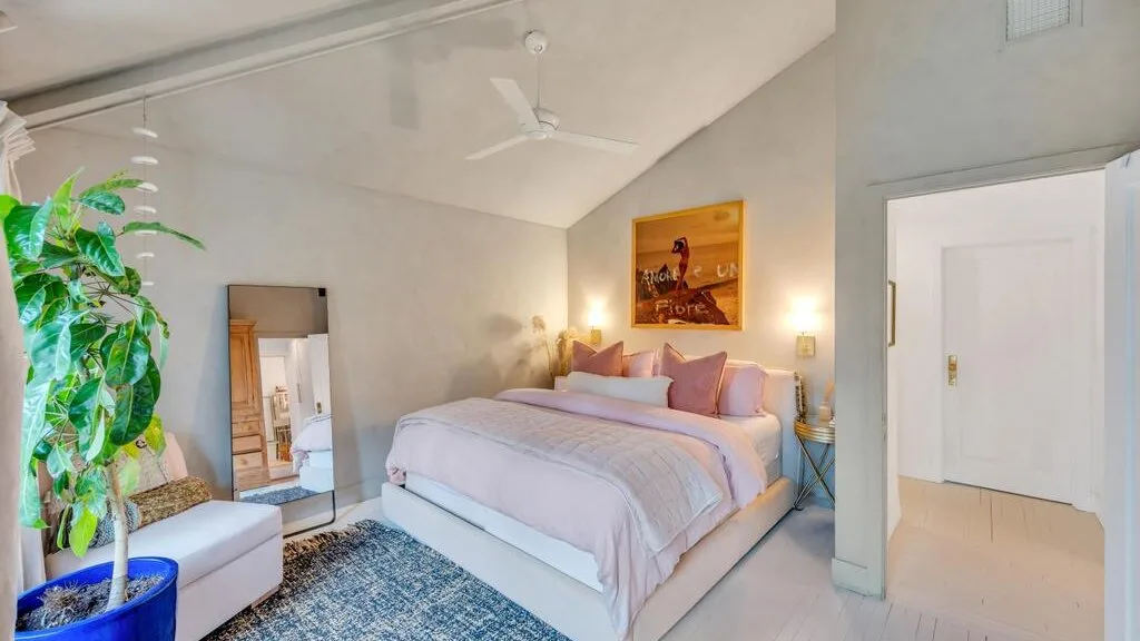 A bedroom with a neatly made bed, bedside lamps, and a large potted plant.