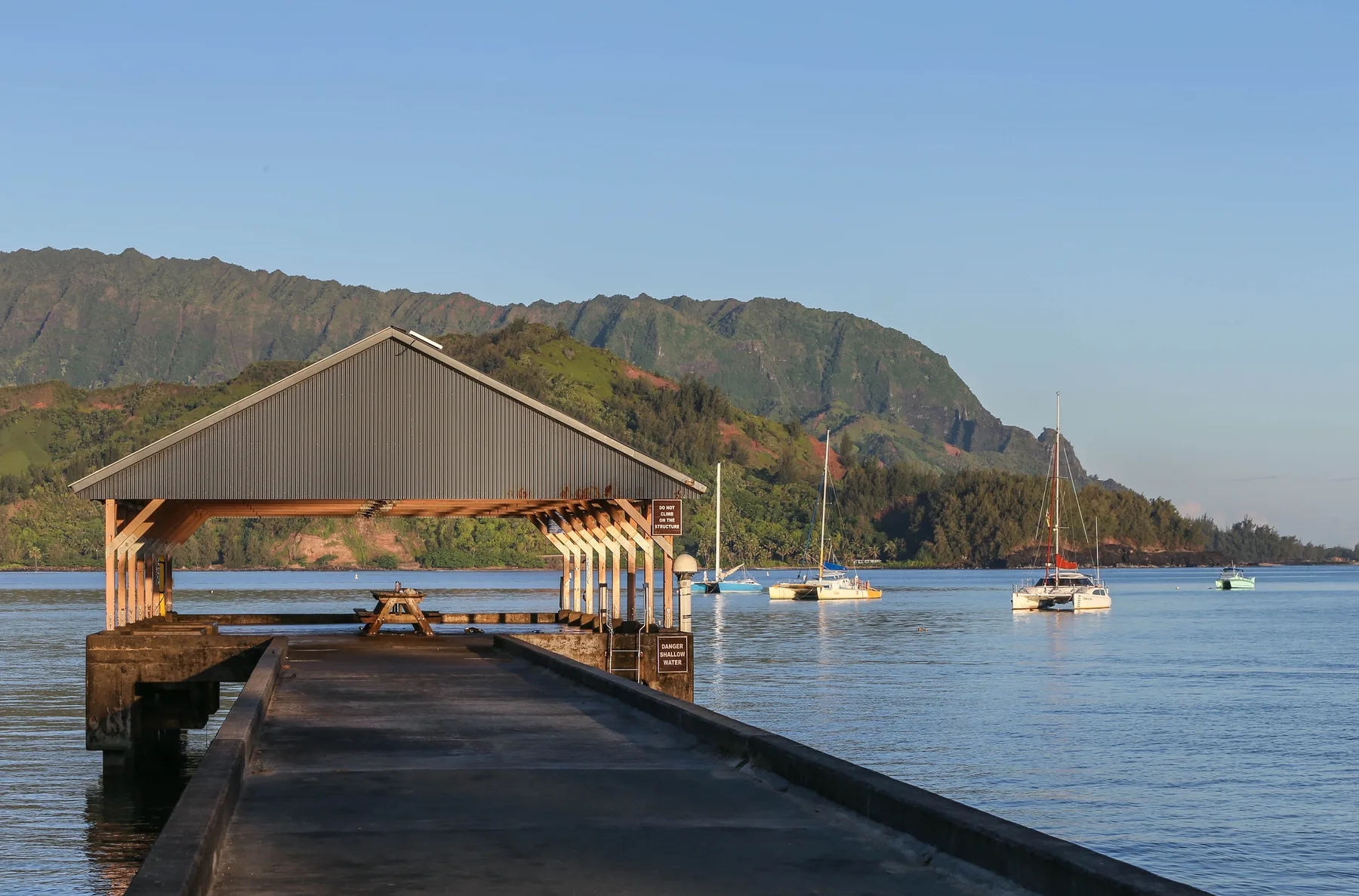Un muelle cubierto se extiende sobre aguas tranquilas, con veleros y una cordillera verde al fondo.