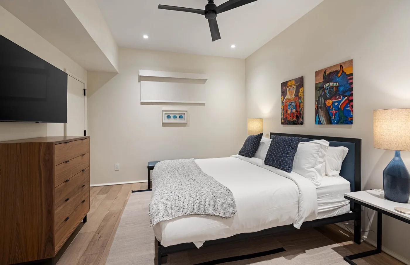 A bedroom with a neatly made bed, colorful wall art, and a ceiling fan.