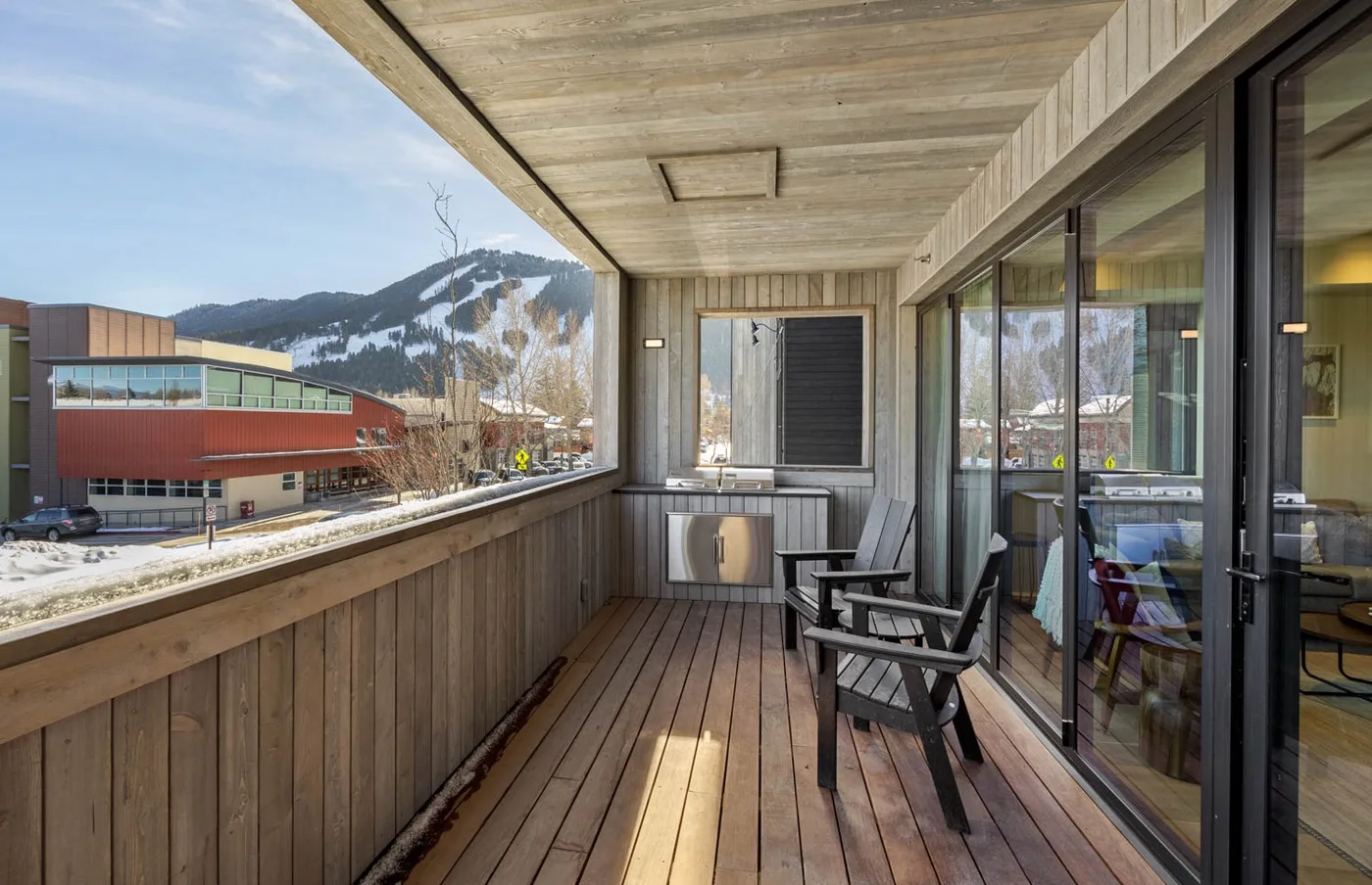 A covered wooden balcony with seating overlooks buildings and a snowy mountain.