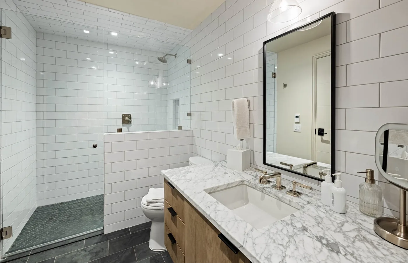 A tiled bathroom with a glass shower, toilet, and a wide vanity with sinks.