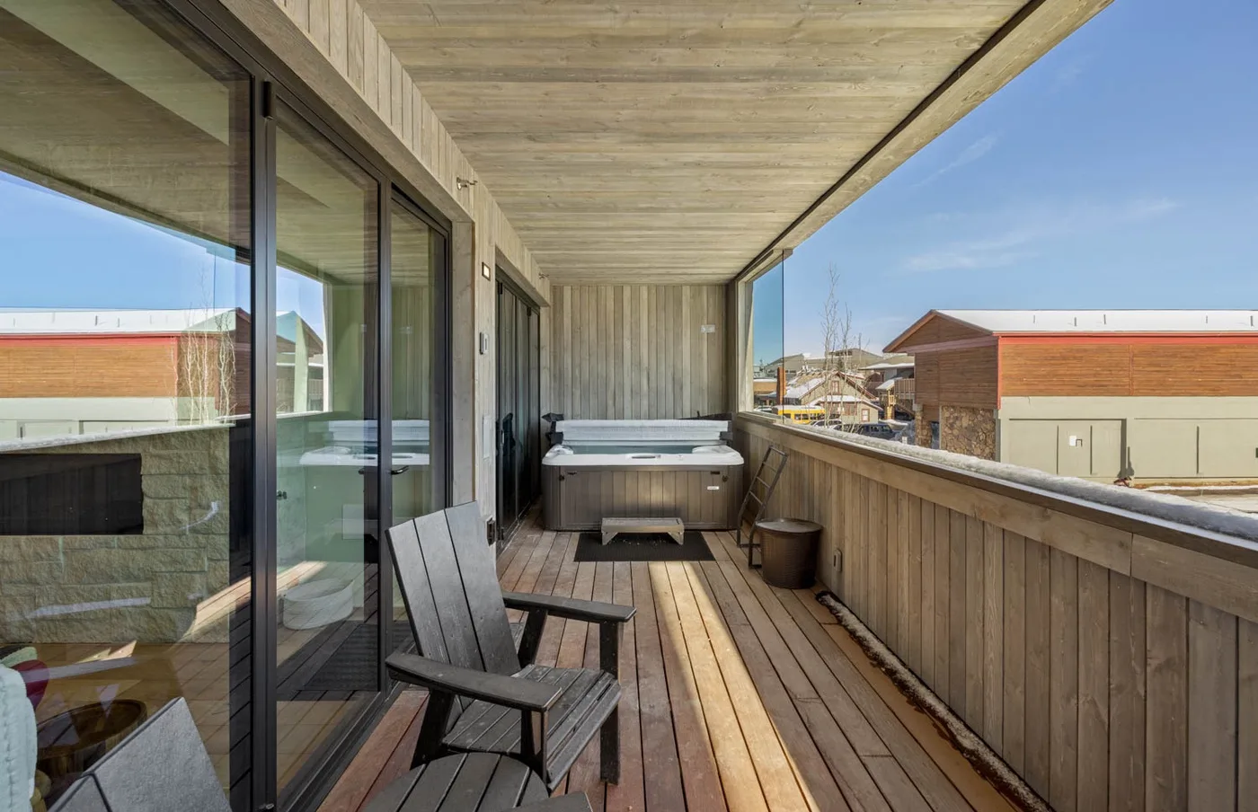 A covered wooden deck has outdoor seating and a hot tub beside glass doors.