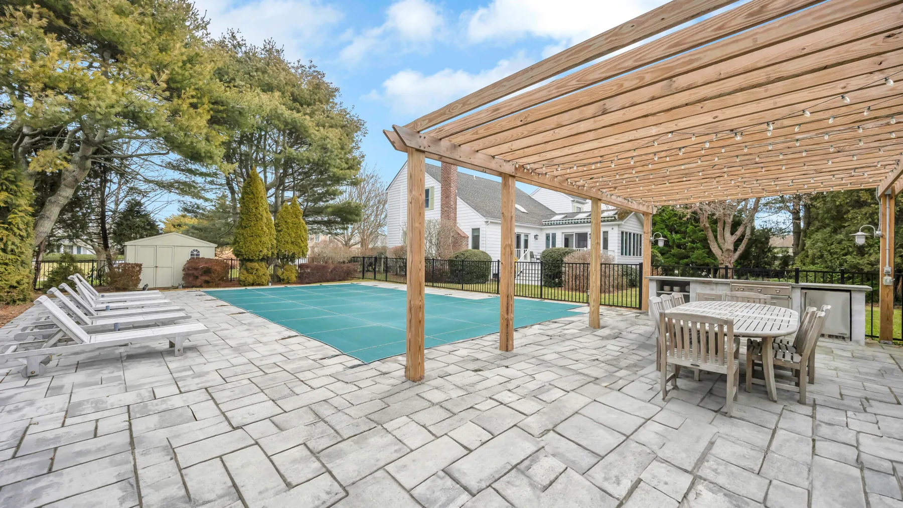 A paved outdoor patio has a swimming pool, lounge chairs, and a wooden pergola.