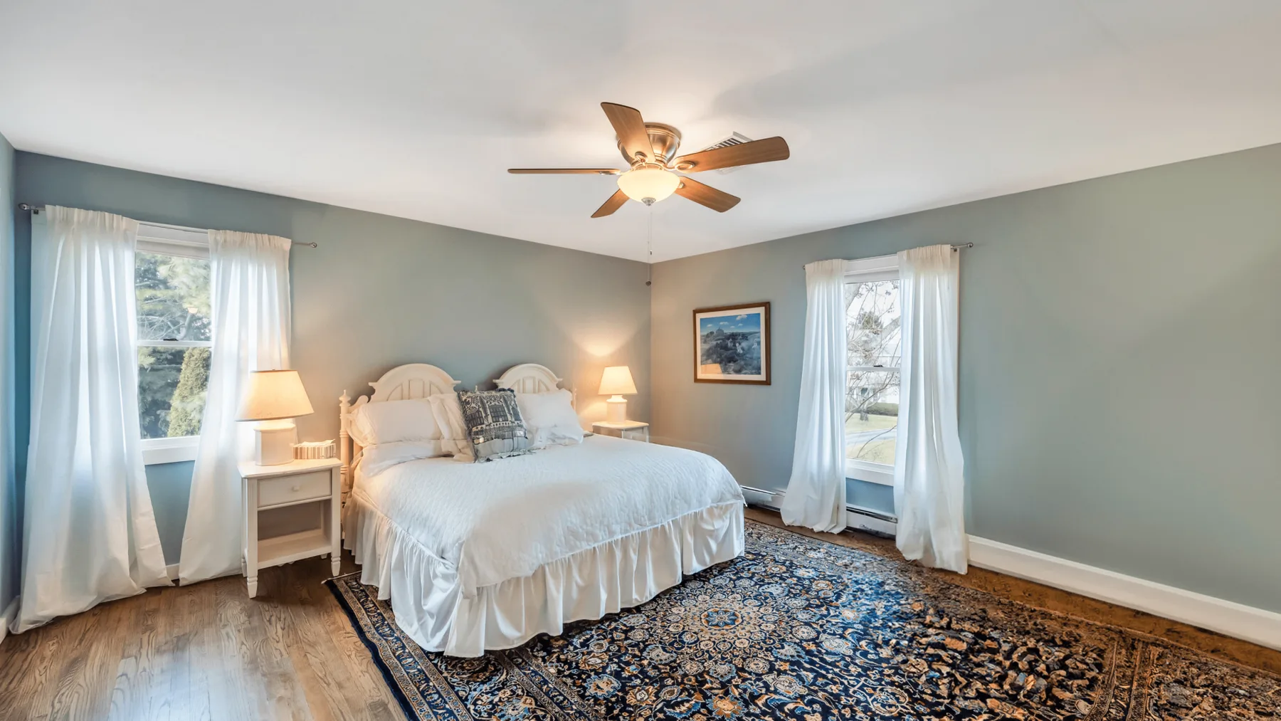 A bedroom with a bed, bedside tables, and windows with light curtains.