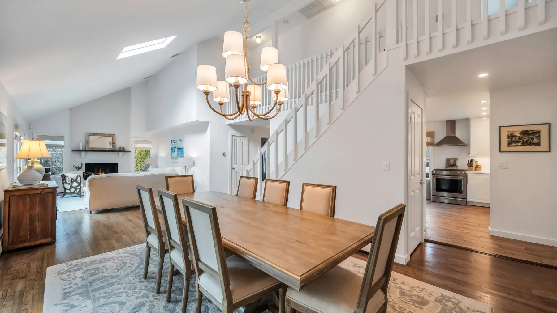 A dining area with a long wooden table beneath a chandelier, beside a staircase.