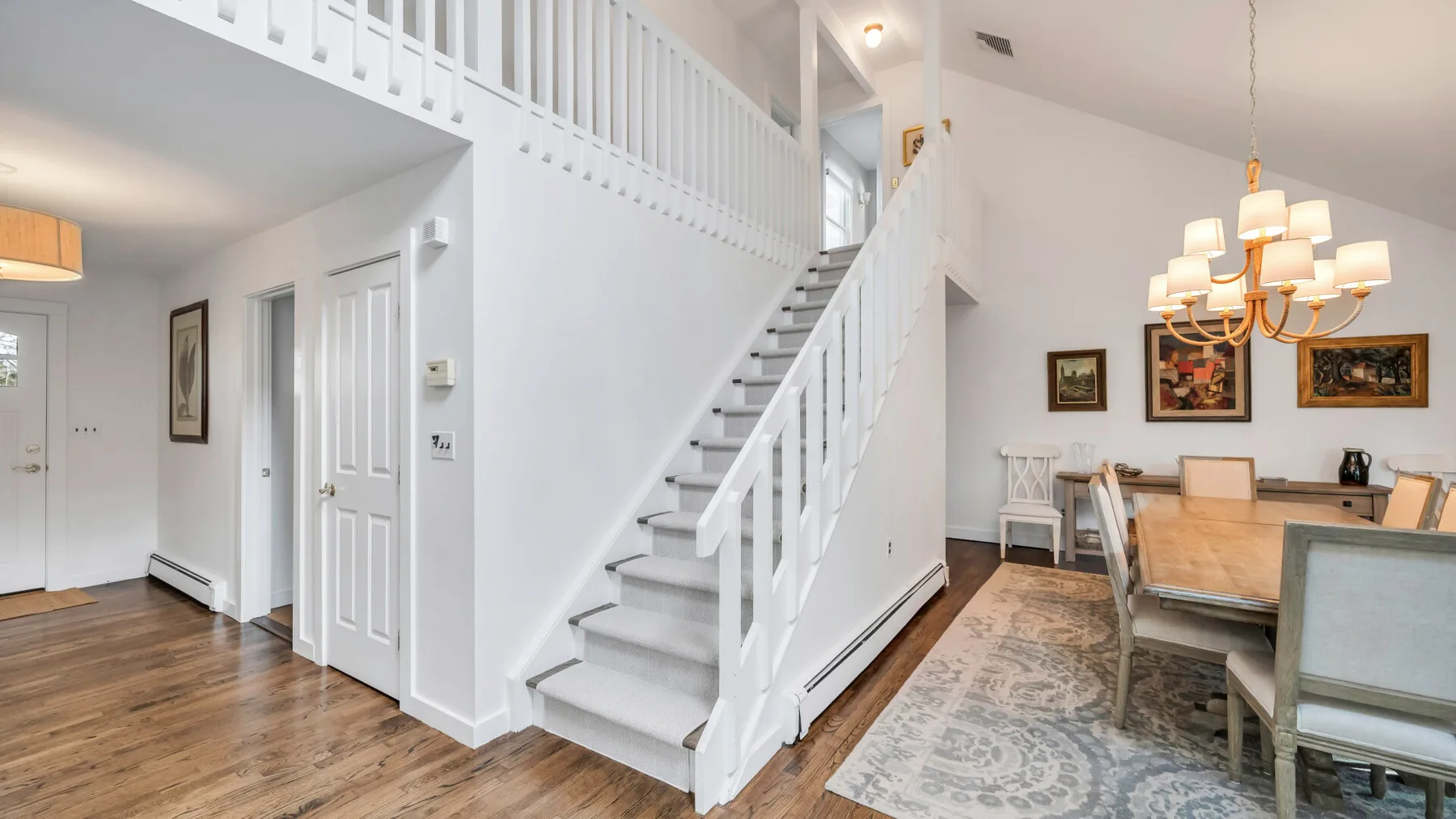A bright dining area beside a white staircase, with a wooden table and chandelier.