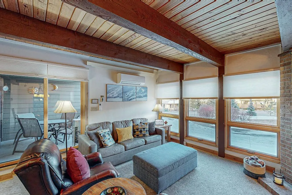 Una sala de estar con asientos grises, grandes ventanas y vigas de madera a la vista.