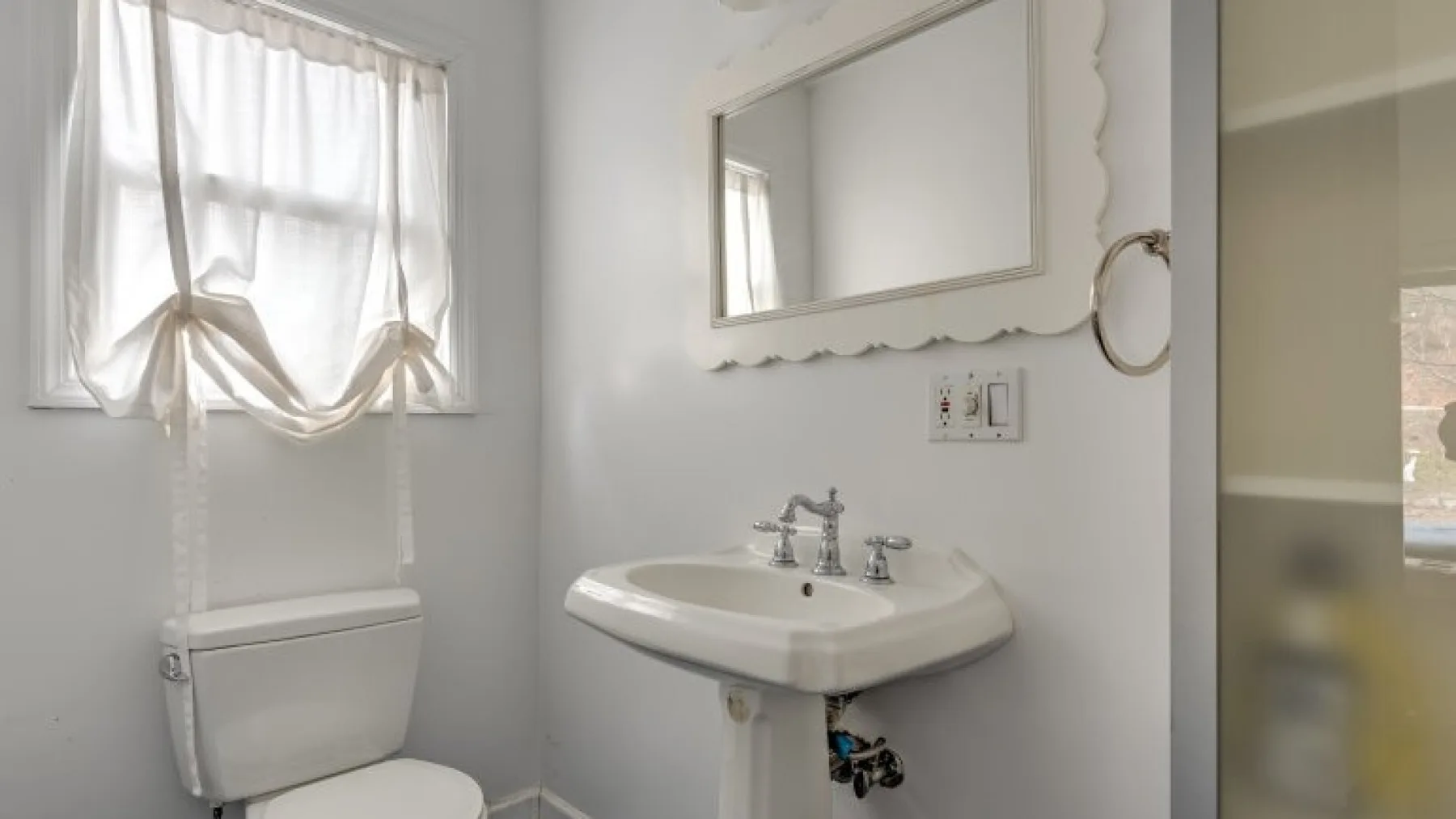 Un baño con lavabo de pedestal, inodoro y una ventana con cortinas translúcidas.