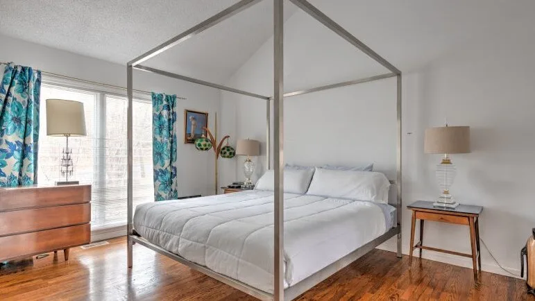 Un dormitorio luminoso con una cama blanca, cortinas estampadas y suelo de madera.
