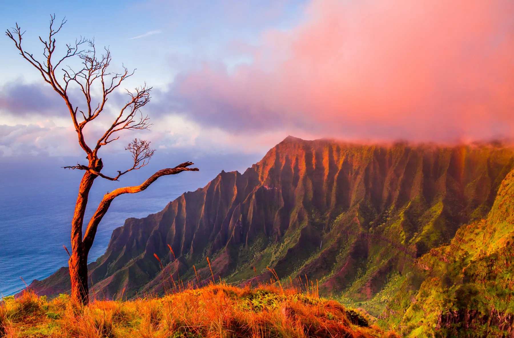 A coastal mountain ridge rises above the ocean beneath colorful clouds.