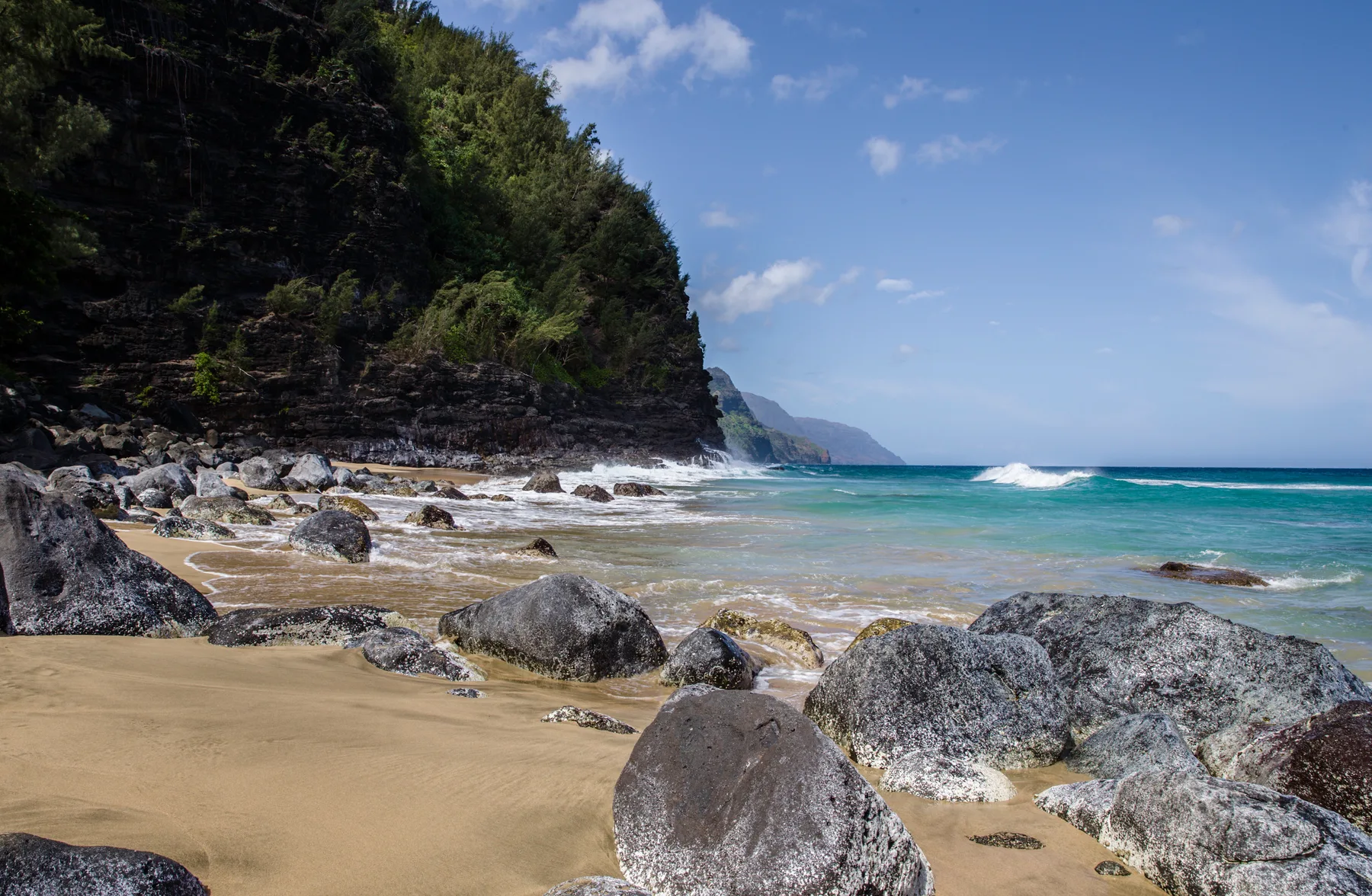A sandy beach with large rocks sits beneath a green coastal cliff.