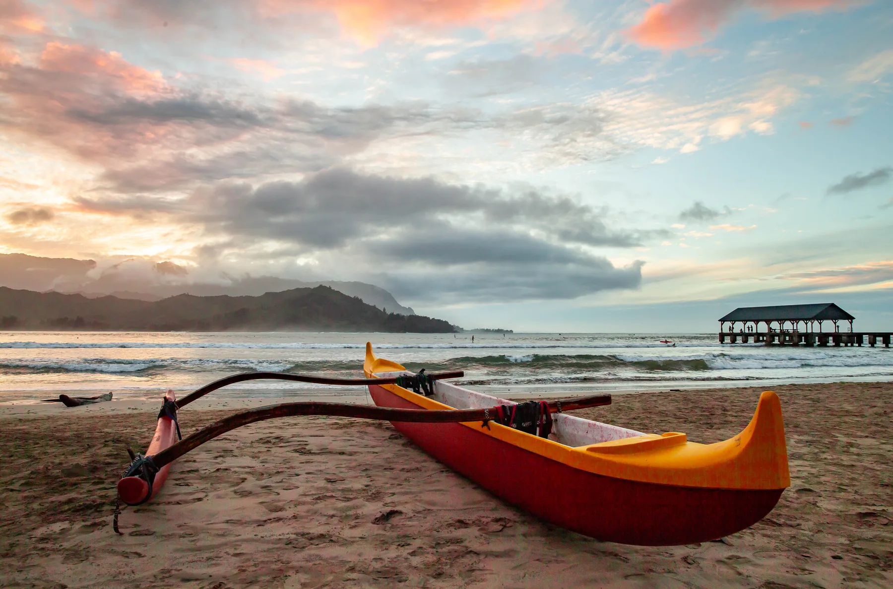 A brightly colored boat rests on a sandy beach beneath a cloudy sunset sky.