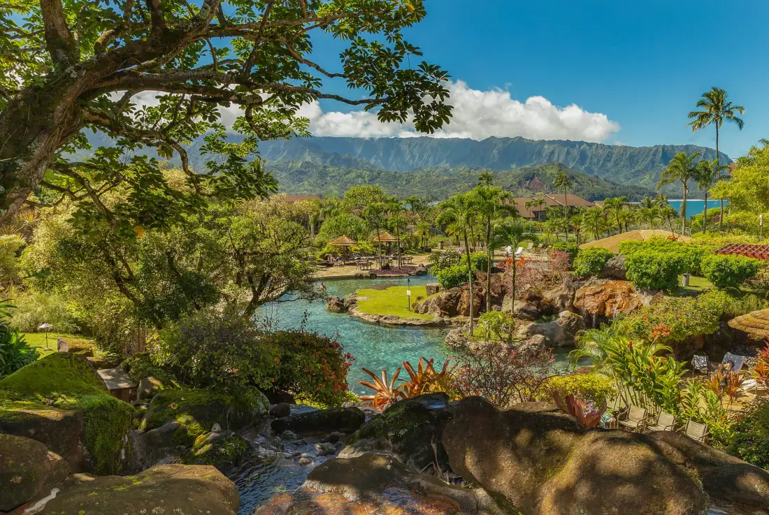 A tropical garden surrounds a turquoise pond beneath a mountain range.