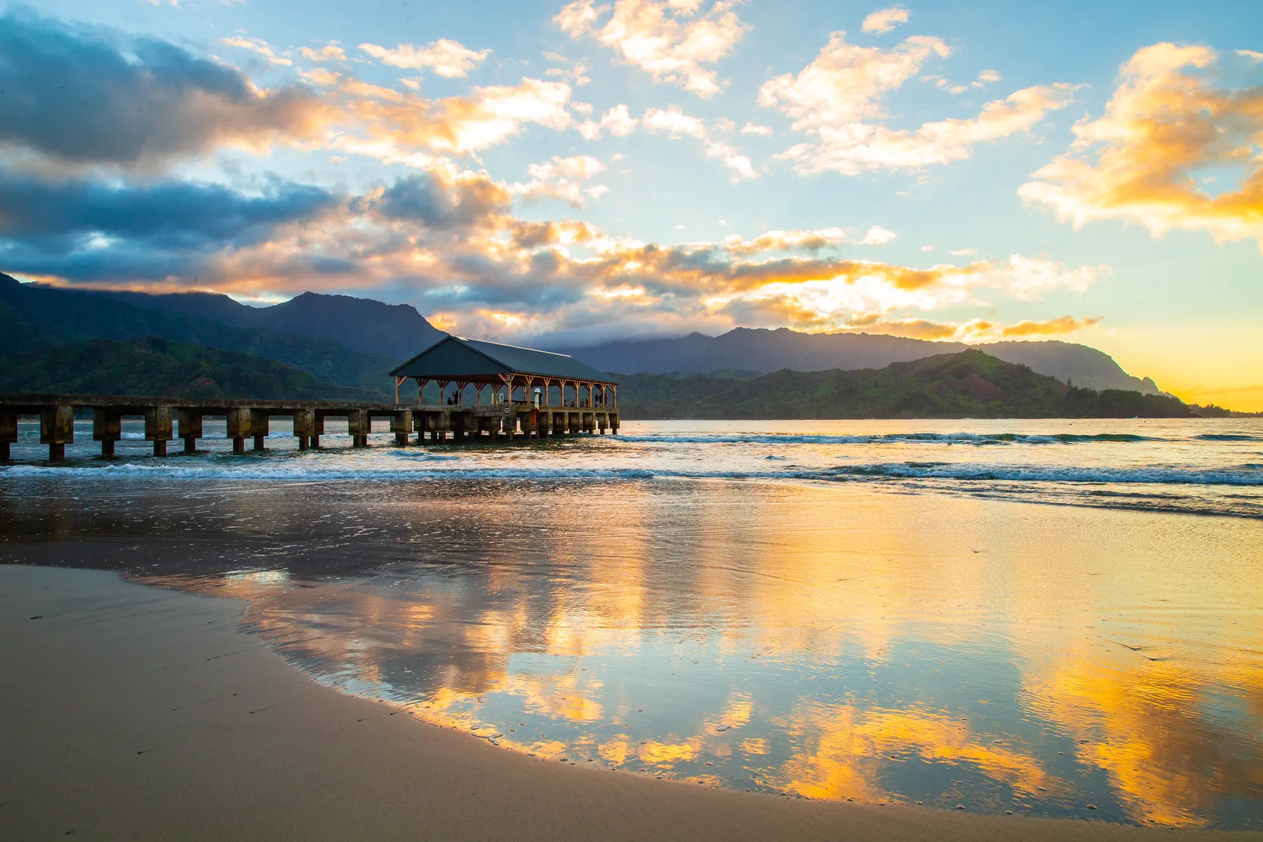 A sunset beach reflects colorful clouds beside a wooden pier and distant mountains.
