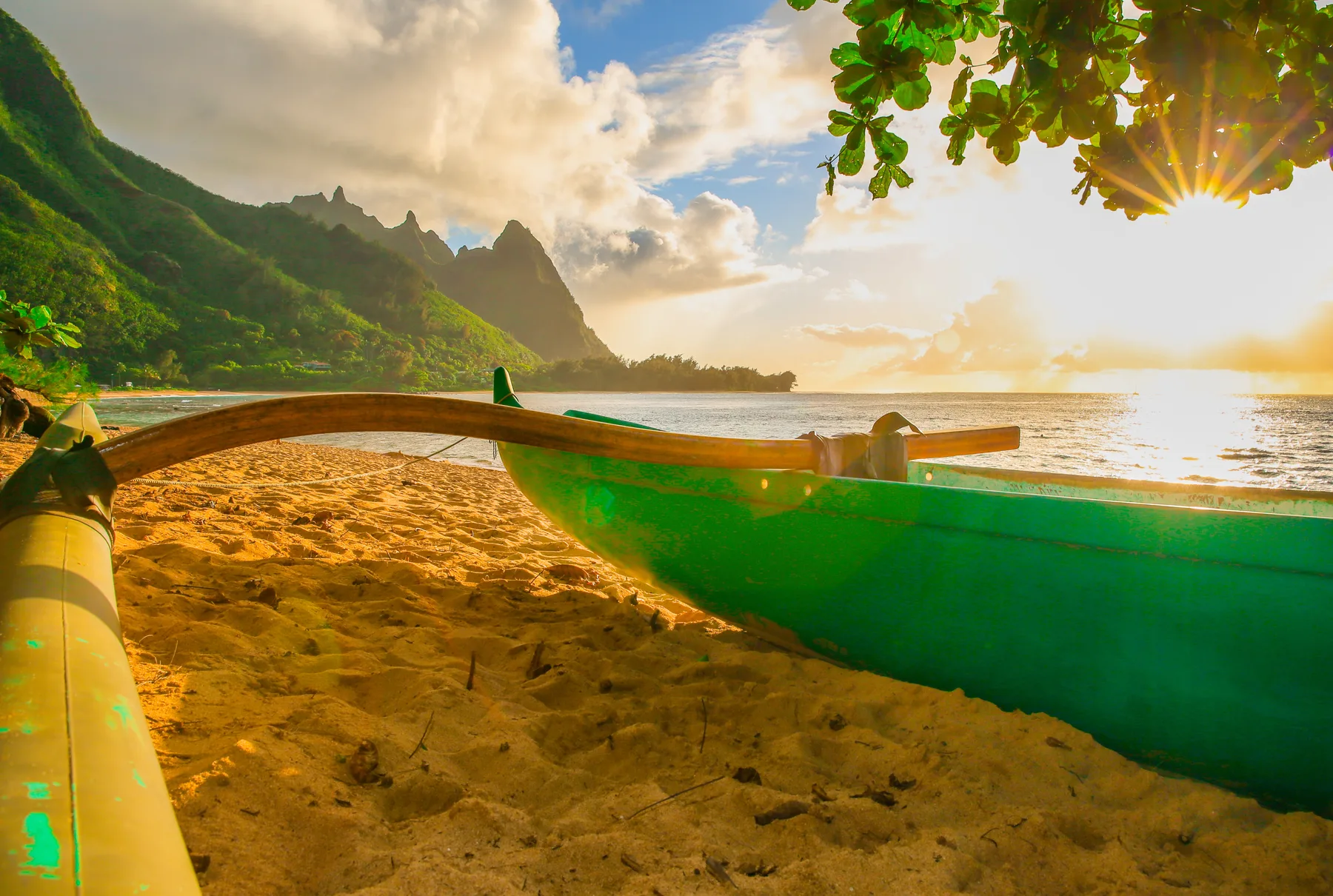 A sandy beach with green boats, coastal mountains, and the sun low over the water.