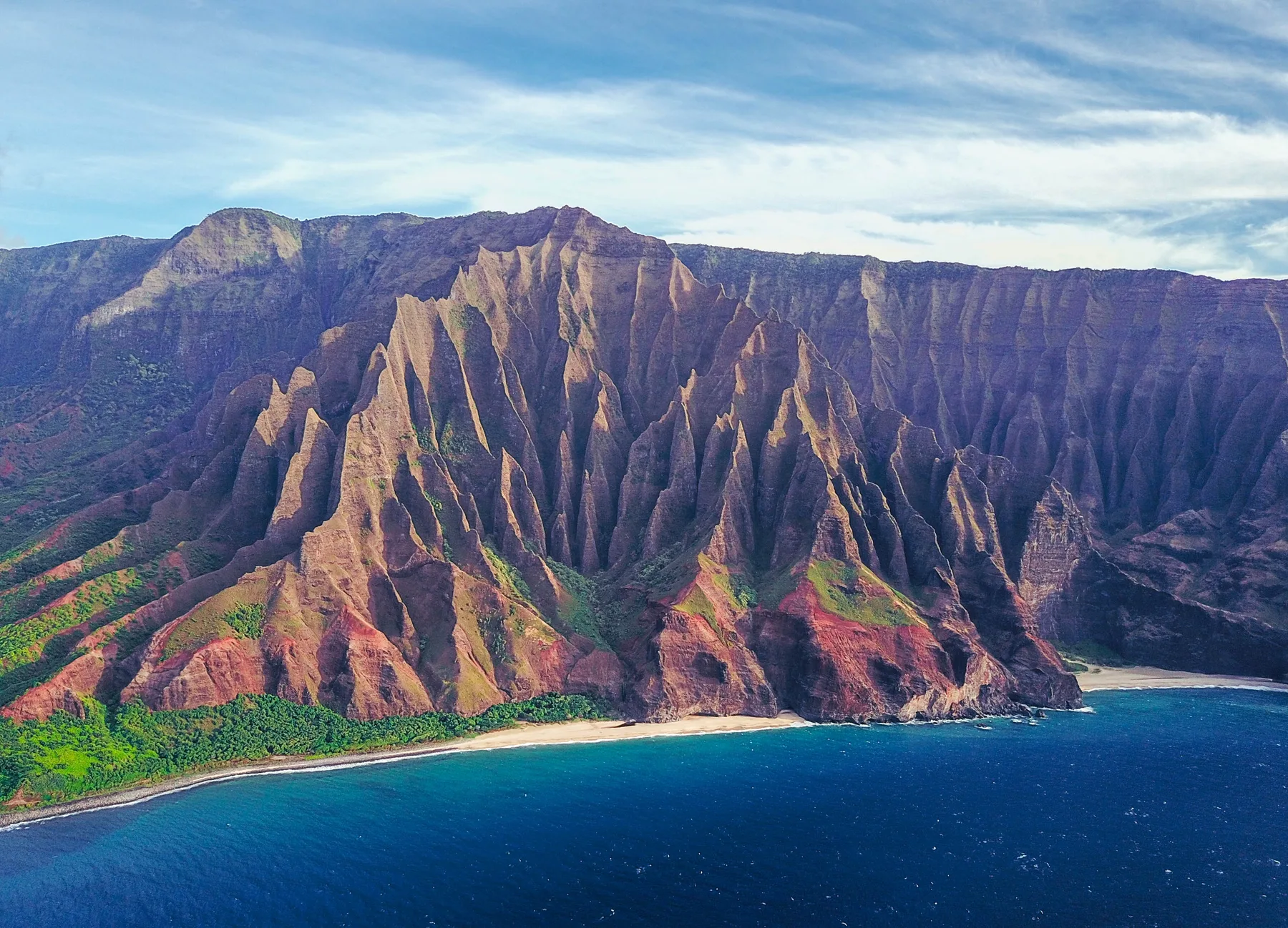 A rugged coastal mountain range rises above a narrow beach and deep blue water.