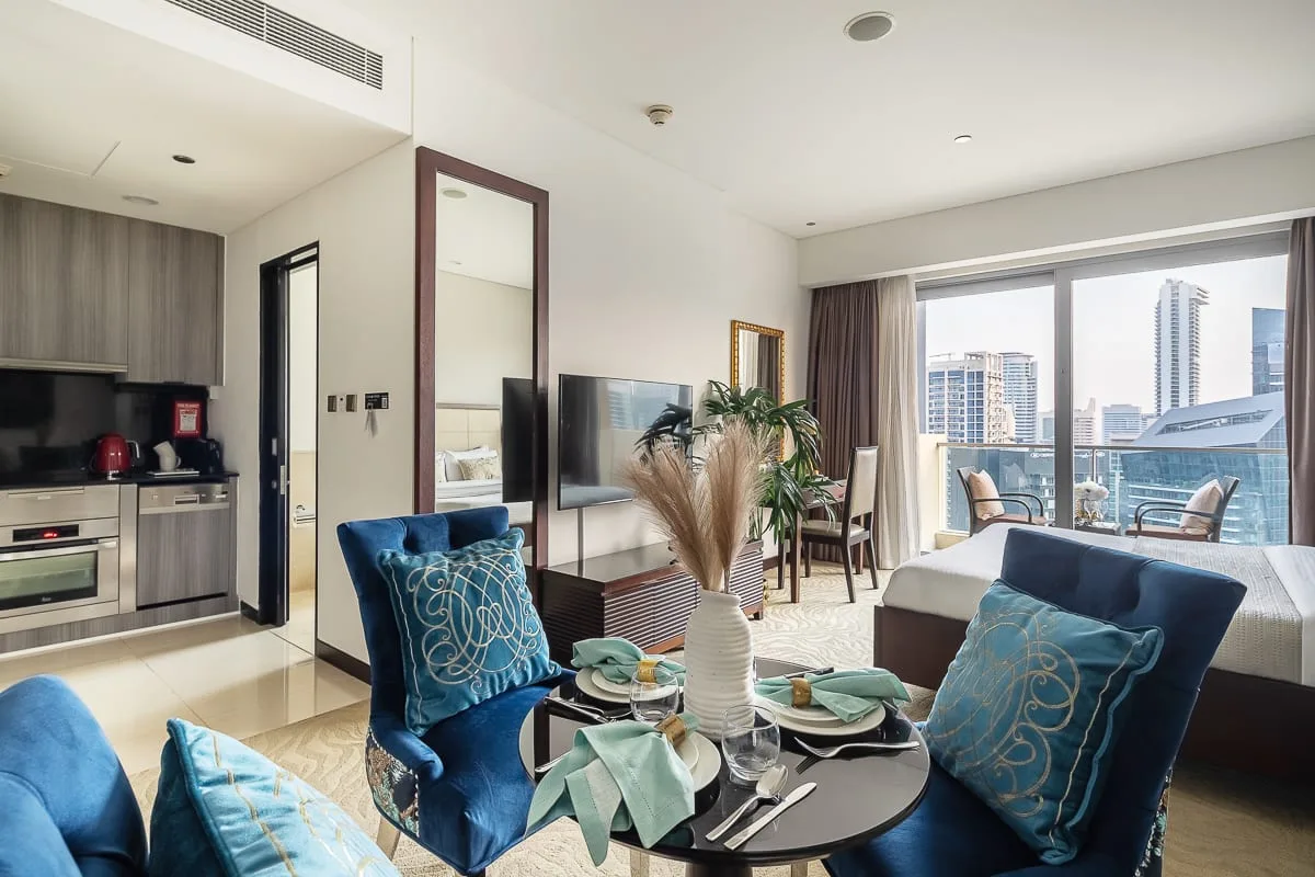 Una sala de estar con asientos azules, un comedor y grandes ventanas con vistas a edificios de la ciudad.