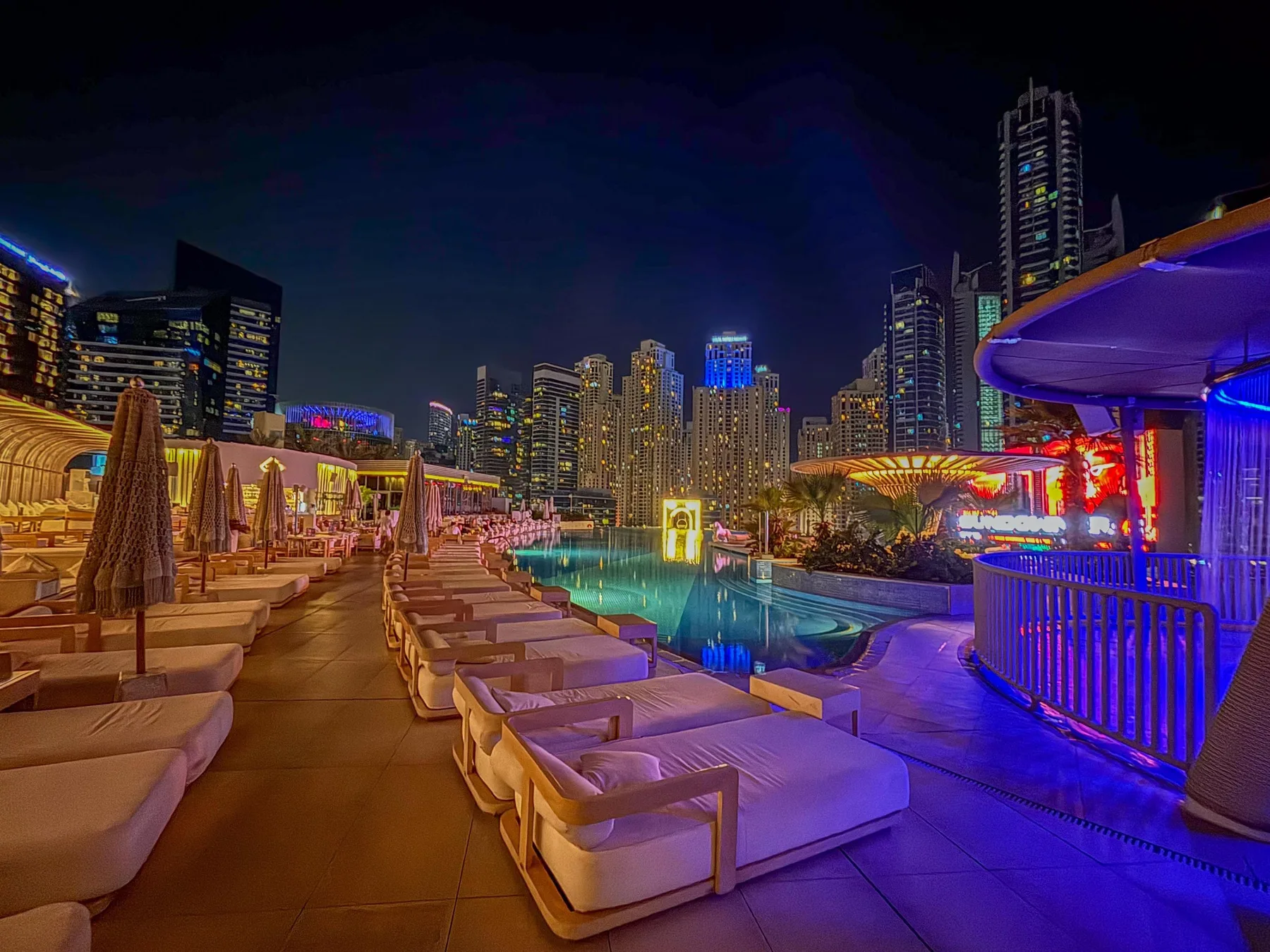A rooftop pool deck at night, lined with lounge seating and city lights.