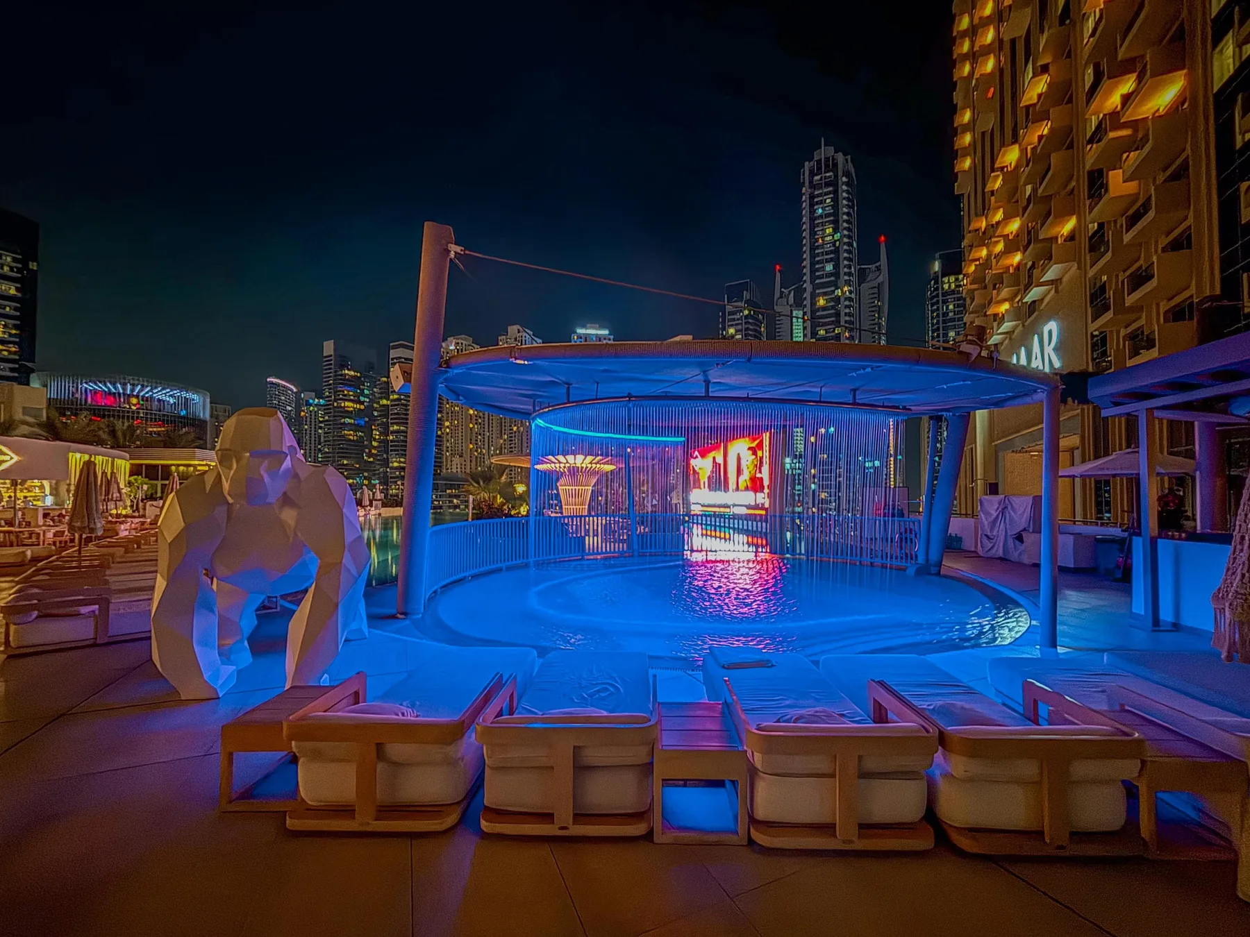 A rooftop pool glows blue at night, with lounge seating and illuminated city buildings.