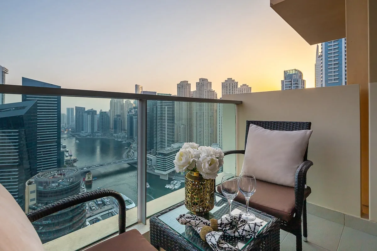 A balcony seating area overlooks a city skyline and waterfront at sunset.