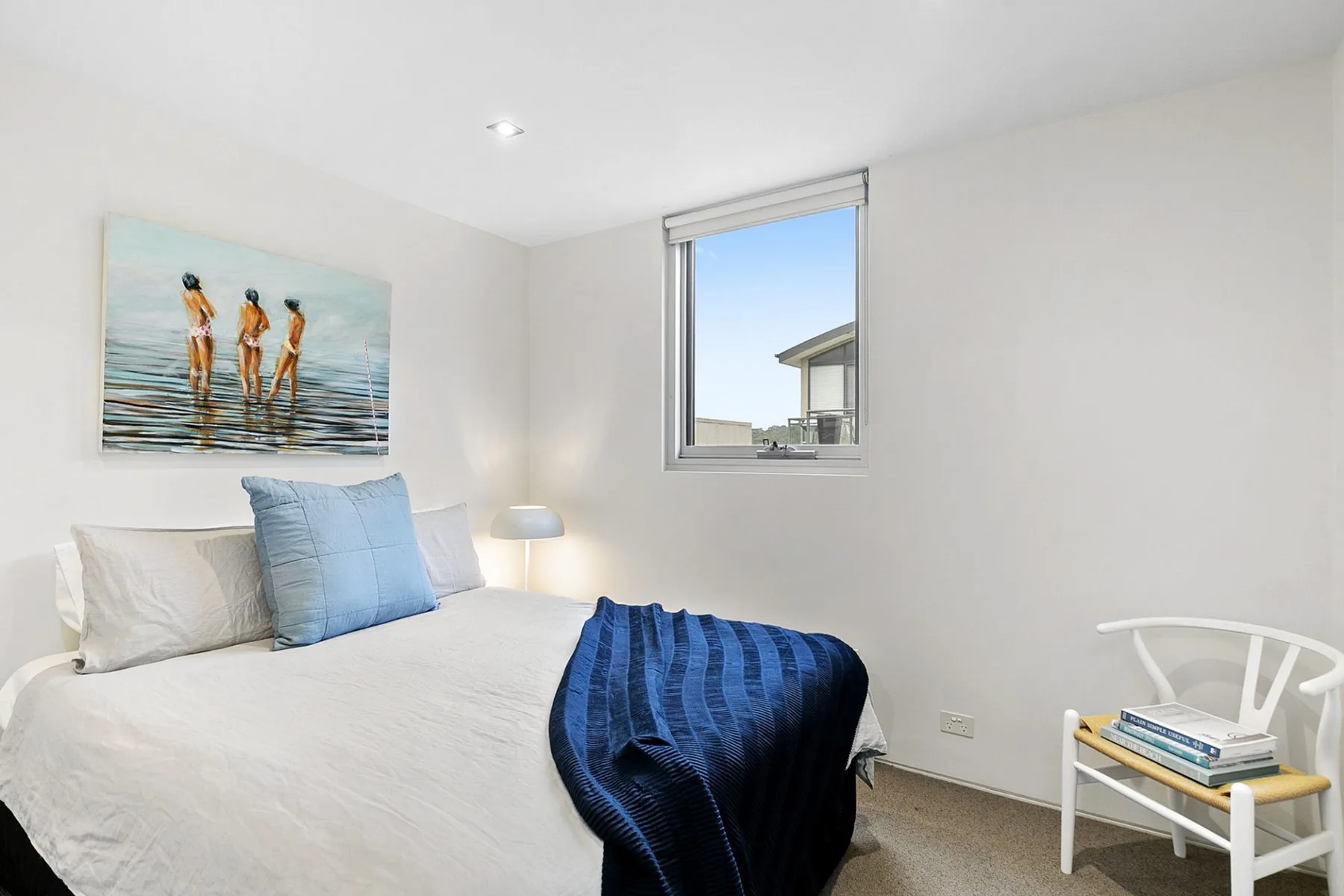 Un dormitorio luminoso con una cama, una manta azul y una silla junto a la ventana.