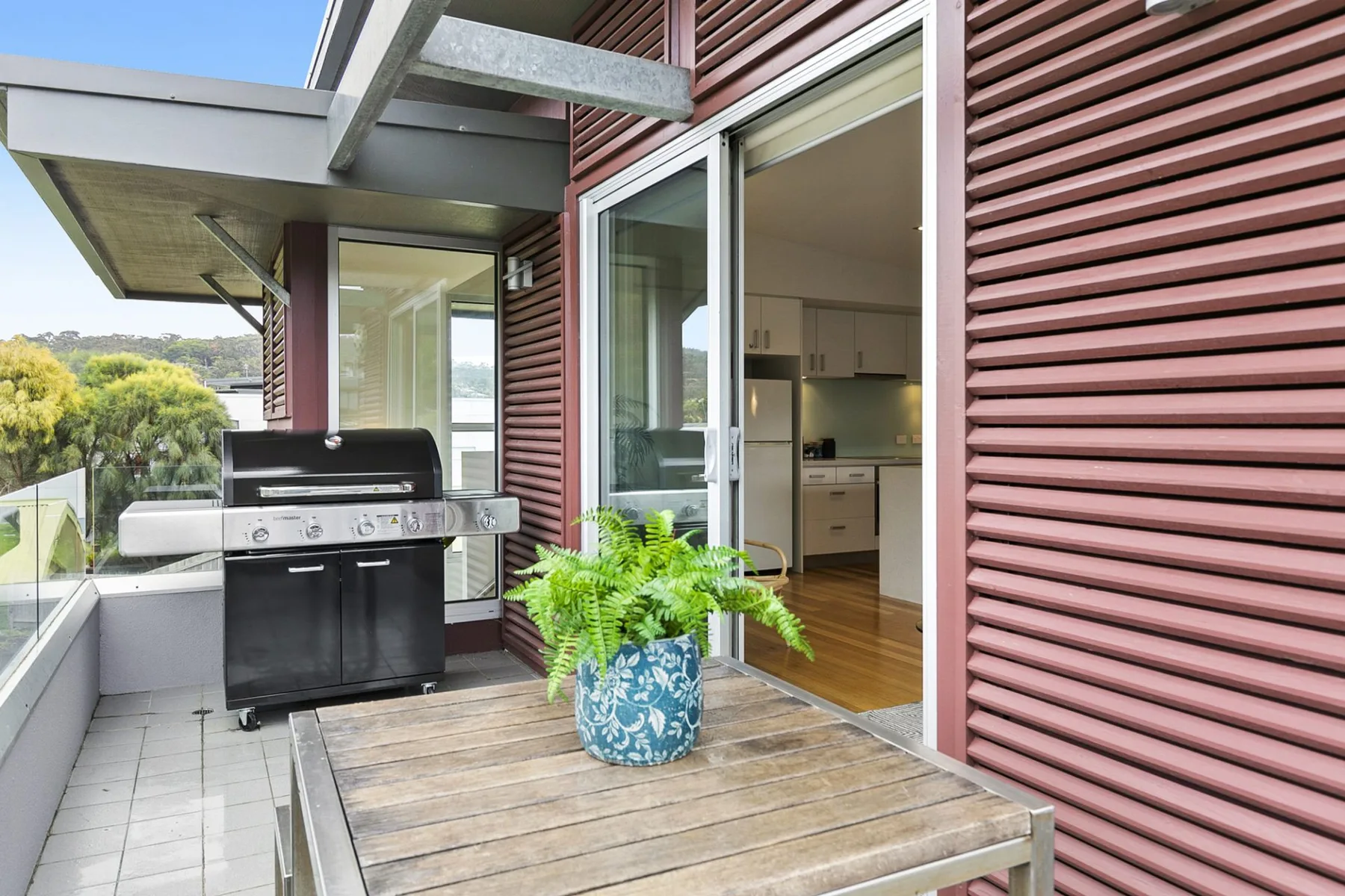 Un patio exterior con una barbacoa, una mesa de madera y un helecho en maceta.