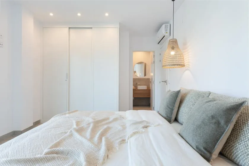 Un dormitorio luminoso con una cama, ropa de cama clara y una lámpara colgante tejida.