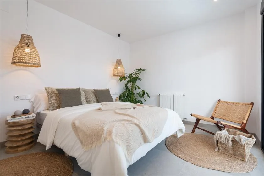 Un dormitorio luminoso con una cama, lámparas colgantes tejidas y una planta frondosa.