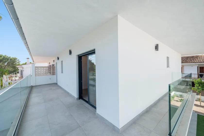 Un balcón con suelo de baldosas recorre una pared exterior blanca con una puerta de vidrio de marco oscuro.